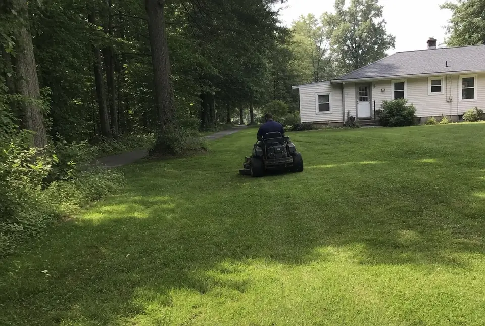 Worker mowing a back lawn beside a wooded trail