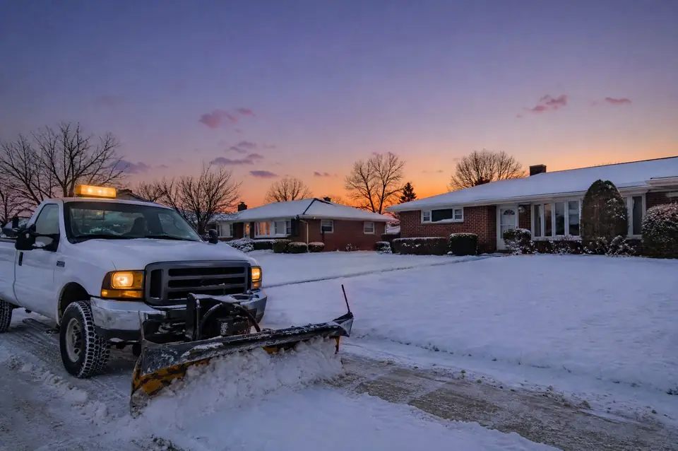 Plow truck clearing a driveway at dusk on a snowy ranch street